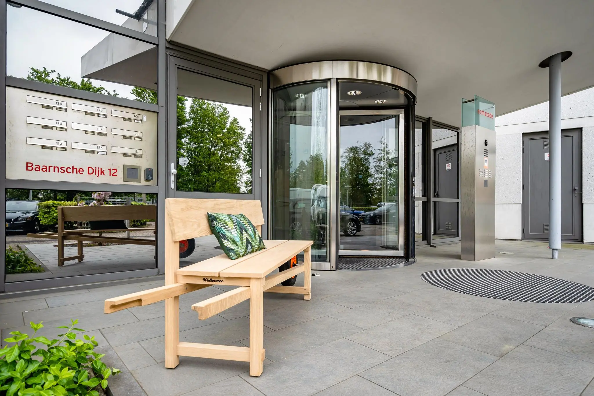 Ingang van een modern kantoorgebouw aan de Baarnsche Dijk 12 met draaideur, houten bank en brievenbussen.